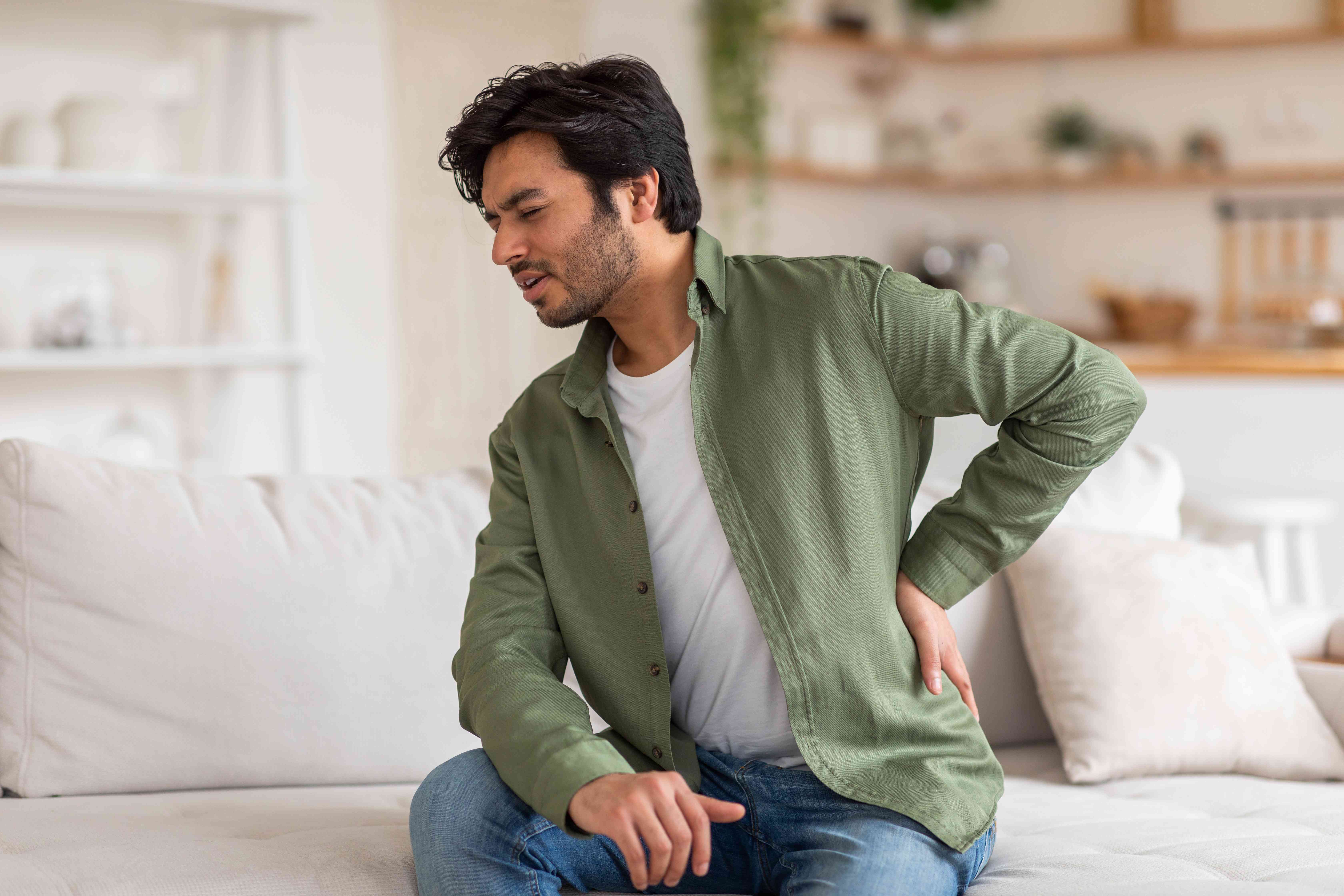 A man sitting on a couch holding his lower back with a pained expression, appearing to suffer from back pain.