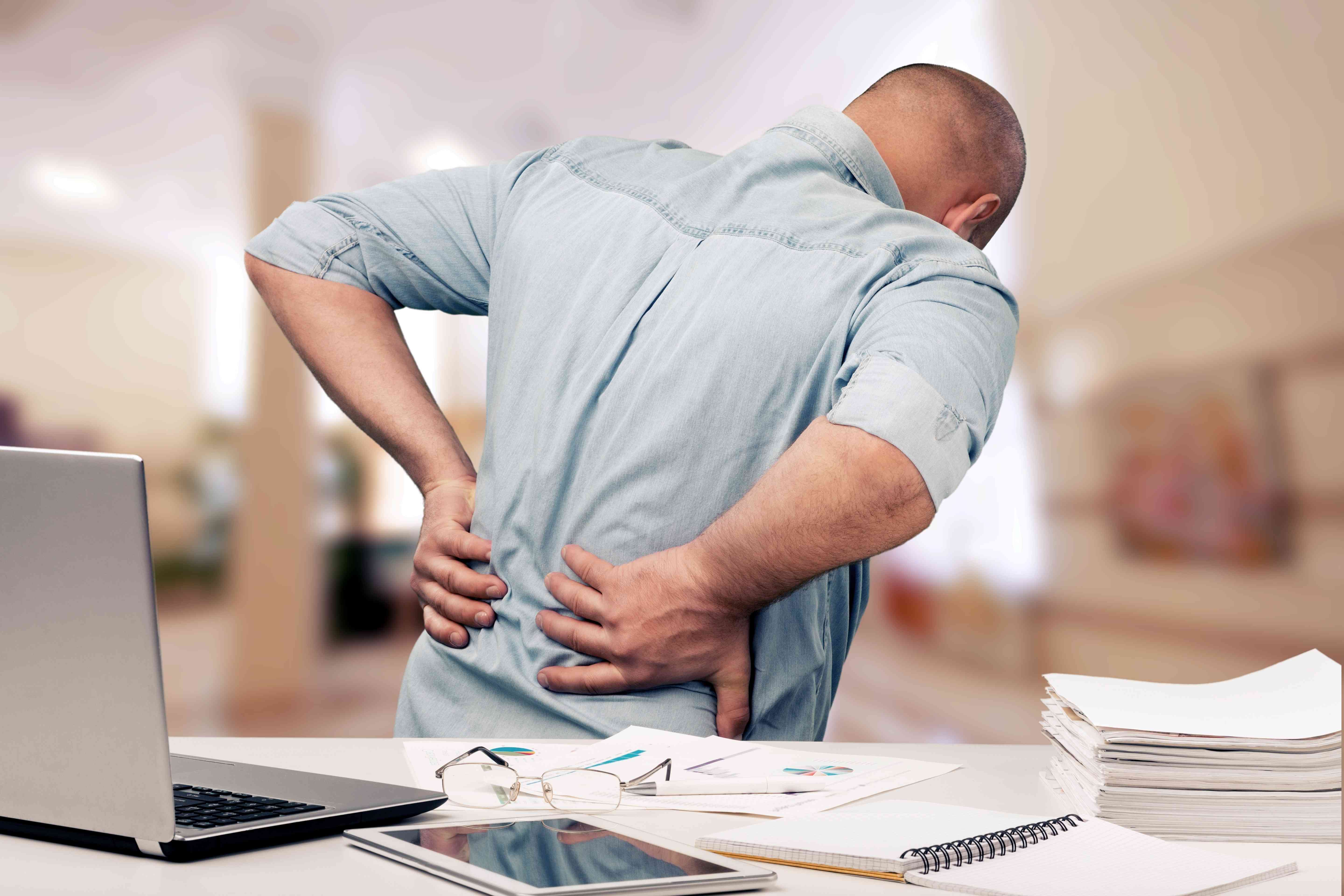 A man standing at his desk with his hands on his lower back, appearing to be in pain, with office supplies and a laptop on the desk.